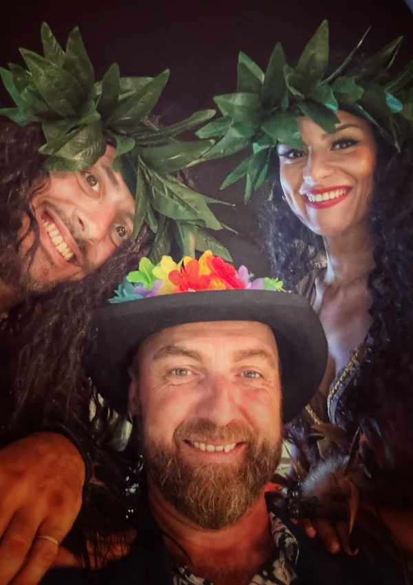magicien-close-up-deambulation-soiree-theme-casino-pleinair-la-ciotat Selfie festif du magicien Zibé arborant un chapeau orné de fleurs colorées aux côtés de danseurs en costumes exotiques avec couronnes de feuilles lors de la Soirée des Îles au Casino Pleinair de La Ciotat.